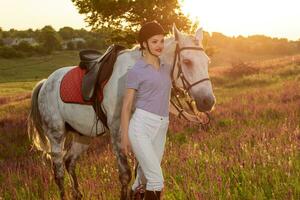 jockey ung flicka petting och kramas vit häst i kväll solnedgång. Sol blossa foto