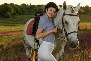 jockey ung flicka petting och kramas vit häst i kväll solnedgång. Sol blossa foto