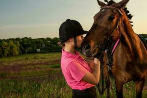 skön leende flicka jockey stå Nästa till henne brun häst bär särskild enhetlig på en himmel och grön fält bakgrund på en solnedgång. foto