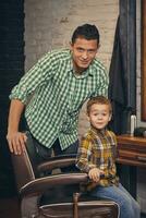 eleganta liten unge Sammanträde på stol på frisör med hans ung far på bakgrund foto