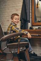 porträtt av en eleganta liten pojke klädd i skjorta och jeans i de frisör, Sammanträde i en stol mot de frisörer arbetsplats foto