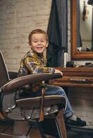 porträtt av en eleganta liten pojke klädd i skjorta och jeans i de frisör, Sammanträde i en stol mot de frisörer arbetsplats foto