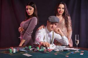 man spelar poker på kasino Sammanträde på tabell med stackar av pommes frites, pengar, kort. fira vinna med två kvinnor. svart bakgrund. närbild. foto