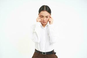 bild av asiatisk serös ser kvinna, finputsning henne huvud med upprörd ansikte uttryck, har huvudvärk, lider från migrän efter påfrestning på arbete, vit bakgrund foto