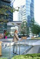 framgångsrik affärskvinna i beige kostym, stående i stad Centrum nära himmel skrotare och kontor byggnader foto
