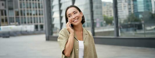 leende ung koreanska flicka talande på mobil telefon och gående i stad. Lycklig kvinna Framställ på gata med smartphone foto