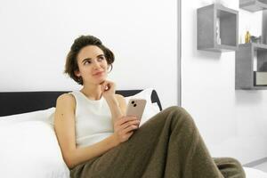 porträtt av ung skön brunett kvinna, Sammanträde på säng med smartphone, använder sig av mobil telefon app, sändning meddelande och leende, läsning på telefon Ansökan i sovrum foto