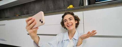 porträtt av ung kvinna video chattar, uppgifter vlog medan sitter på kök golv. söt flicka tar selfie på smartphone foto