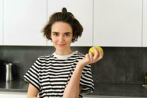 porträtt av ung kvinna modell, Framställ i de kök, innehav ett äpple, leende och ser på kamera foto