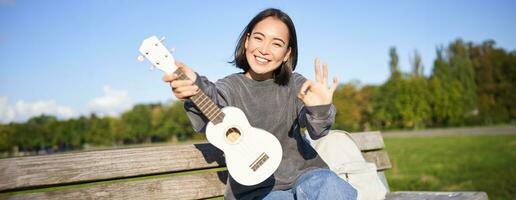 söt leende flicka visar ok tecken och henne ny ukulele, sitter på bänk i parkera, rekommenderar musikalisk instrument foto