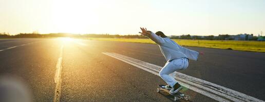 Lycklig skater flicka ridning henne skateboard och har roligt på tömma gata. leende kvinna njuter kryssare rida på solig väg foto