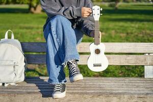 beskurna skott av ung flicka i gymnastikskor och jeans, händer innehav ukulele musikalisk instrument medan hon sitter på bänk i grön solig parkera foto