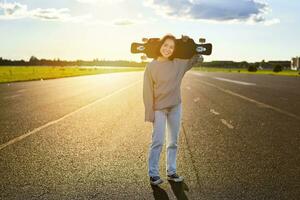 asiatisk flicka med skateboard stående på väg under solnedgång. skater Framställ med henne lång styrelse, kryssare däck under Träning foto