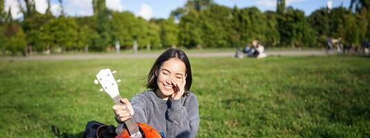 musik och instrument. leende asiatisk flicka visar henne vit ukulele, sitter i parkera och spelar små gitarr foto