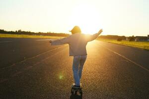 tonåring flicka känsla Lycklig på longboard. Lycklig ung skater ridning henne skateboard med händer spridning åt sidan, känsla frihet, gående mot de Sol foto