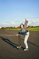 vertikal porträtt av leende asiatisk kvinna stående på väg med longboard, skateboard på lång kryssare, Framställ på tömma väg på solig dag foto