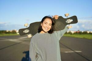 fritid och människor. Lycklig asiatisk kvinna stående med longboard, cruising på ett tömma väg i landsbygden. skater flicka innehar henne skateboard och ler på kamera foto