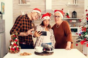 niece fira jul med morförälder framställning rolig ansikten medan tar selfies. Lycklig glad mång generation familj använder sig av telefon till ta Foto bär santa hatt håller på med hand gest med jul träd i de bakgrund.