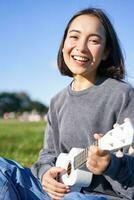 vertikal skott av Lycklig koreanska flicka Sammanträde i parkera, inlärning på vilket sätt till spela ukulele, sång och avkopplande foto