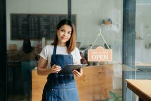 ung kvinna hängande en Välkommen tecken i främre av en kaffe affär. skön servitris eller värdinna innehav en läsplatta framställning i en restaurang. foto