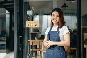 ung kvinna hängande en Välkommen tecken i främre av en kaffe affär. skön servitris eller värdinna innehav en läsplatta framställning i en restaurang. foto