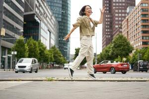 porträtt av asiatisk Lycklig flicka Hoppar och dans i stad Centrum, Framställ på gator, uttrycka glädje och spänning foto