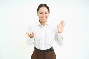 porträtt av skön asiatisk kvinna höjer ett hand, ler vänlig, introducerar själv, hälsar och säger Hallå, vit bakgrund foto
