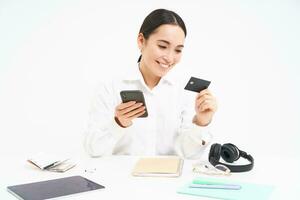företags- och företag. asiatisk kvinna i kontor, innehar smartphone och kreditera kort, betalande uppkopplad, miljö upp direkt debitera uppkopplad, vit bakgrund foto