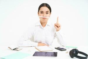 företag och kontor arbete. ung kvinna Sammanträde på arbetsplats, lurar runt om, innehar penna med mun och visar rolig ansikten, vit bakgrund foto