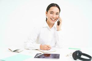 företag och företag begrepp. ung asiatisk kvinna, chef arbetssätt i kontor, skrivning ner information och talande med klient på mobil telefon, vit bakgrund foto