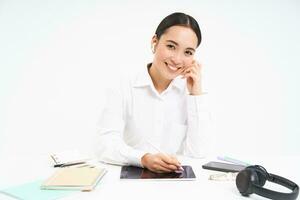 porträtt av asiatisk kvinna i hörlurar, arbetssätt på digital läsplatta, signering digital kontrakt, Sammanträde i kontor och leende, vit bakgrund foto