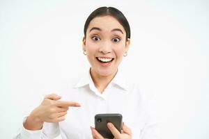 stänga upp porträtt av Lycklig försäljare, framgångsrik kvinna med smartphone, pekande på henne telefon och leende förvånad, stående över vit bakgrund foto