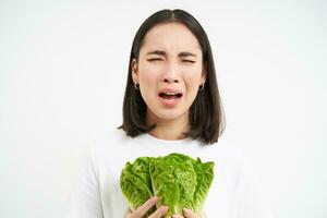 friska diet och rå mat. ledsen och olycklig asiatisk kvinna, innehav färsk sallad löv, kål i händer, äter grönsaker, vit bakgrund foto
