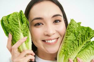 friska mat och vegan livsstil. stänga upp porträtt av leende asiatisk kvinna, utseende Lycklig, visar henne ansikte med sallad, äter kål, vit bakgrund foto