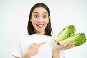 porträtt av överraskad och förvånad asiatisk kvinna, poäng på kål, hitta ut Fantastisk fördel av sallad, friska grönsaker, vit bakgrund foto