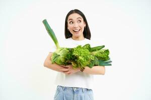 porträtt av leende koreanska kvinna, kramar henne grönsaker, gillar äter rå mat, vegetarian åtnjuter näringsrik diet, vit bakgrund foto