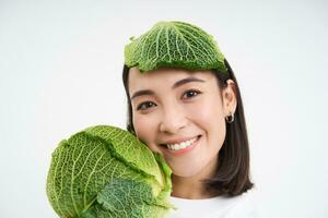 stänga upp porträtt av söt asiatisk kvinna med sallad blad på huvud, leende och som visar grön kål, vit bakgrund foto