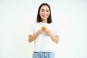 porträtt av koreanska kvinna, som visar cupcake, äter kaka och leende, isolerat på vit bakgrund foto