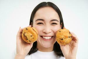 bild av koreanska kvinna åtnjuter äter bakad bakverk, som visar två gott muffins nära ansikte och ler, vit bakgrund foto