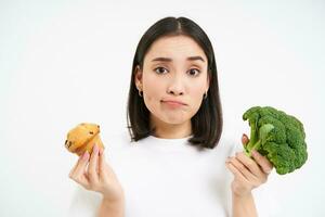 kvinna med ledsen ansikte, innehav vegetabiliska och bakverk, upprörd varelse på diet, välja broccoli istället av cupcake, vit bakgrund foto