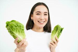 leende asiatisk kvinna, vegan äter kål, åtnjuter gott färsk sallad löv, står över vit bakgrund foto