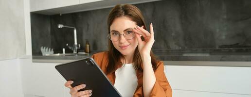 porträtt av skön ung kvinna modell i glasögon, innehav digital läsplatta, bär glasögon, läsning e-bok, Framställ i kök på Hem foto
