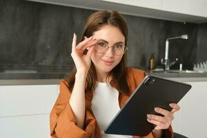 porträtt av skön ung kvinna modell i glasögon, innehav digital läsplatta, bär glasögon, läsning e-bok, Framställ i kök på Hem foto