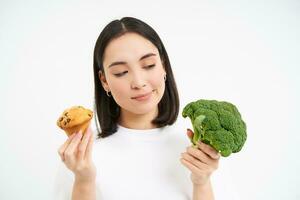 porträtt av asiatisk flicka tänkande Vad till äta, välja mellan muffin och broccoli grönsaker, vit bakgrund foto