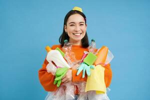 porträtt av leende koreanska kvinna, innehav tömma plast flaskor, sopor för återvinning, ser entusiastisk, sortering ut skräp, blå bakgrund foto