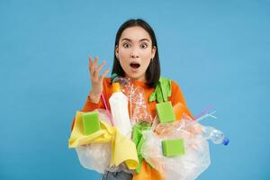 asiatisk kvinna utseende chockad, innehar plast skräp, sortering sopor för återvinning, utseende överraskad, blå bakgrund foto