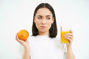 stänga upp porträtt av ung osäker flicka, innehav orange frukt och ser tveksam på glas av juice, står över vit bakgrund foto