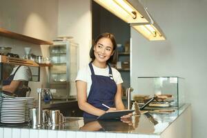 porträtt av skön asiatisk flicka leende, Barista i Kafé arbetssätt Bakom disken, använder sig av läsplatta som pos terminal, bearbetning beställa foto