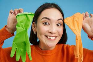 stänga upp porträtt av leende asiatisk kvinna, som visar två annorlunda latex handskar för rengöring, ser entusiastisk, håller på med hus ärenden, blå bakgrund foto