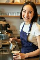 vertikal skott av vänlig Barista flicka, leende på du medan framställning kaffe, häller mjölk och framställning cappuccino foto